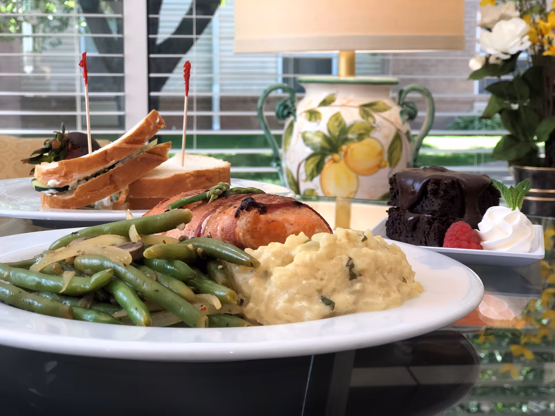A close-up view of a meal on a glass table featuring green beans with onions, mashed potatoes, and a piece of bacon-wrapped salmon. In the background, there is a plate with two sandwiches held together with toothpicks, and a dessert plate with a chocolate cake, whipped cream, a raspberry, and a mint leaf. Behind the food, there is a decorative lamp with a lemon design and a vase with flowers near a window with horizontal blinds.