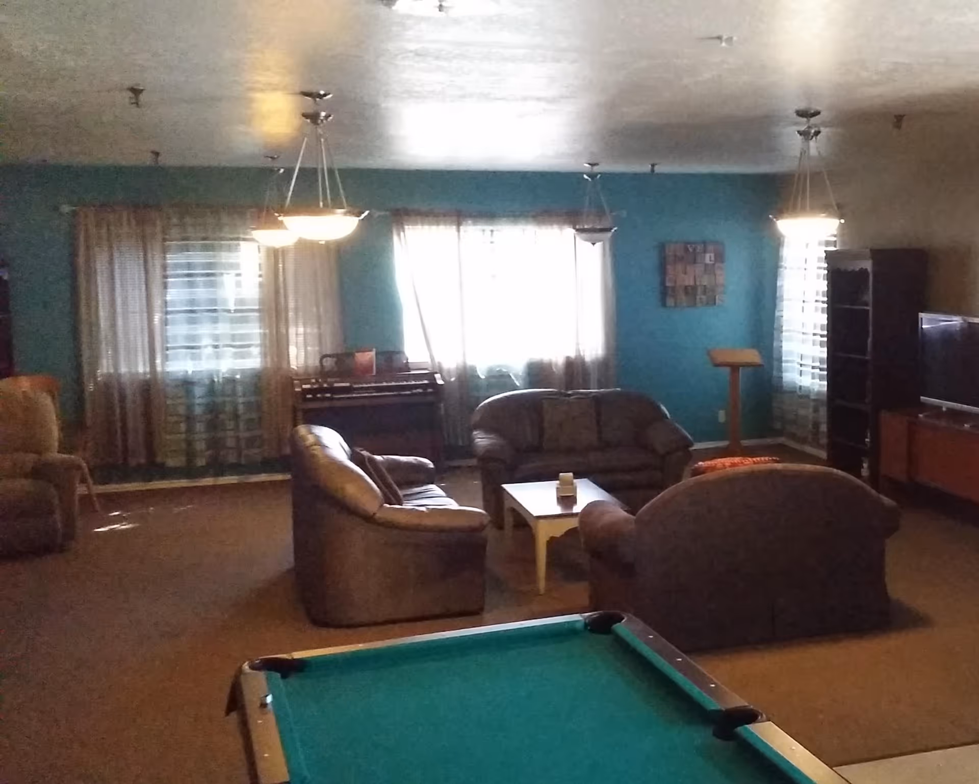 A common lounge area with several brown cushioned sofas arranged around a white coffee table. There is a pool table in the foreground, a television on a stand to the right, and a piano against the back wall. The walls are painted teal, and there are three windows with sheer curtains letting in natural light. Ceiling lights hang above the seating area.