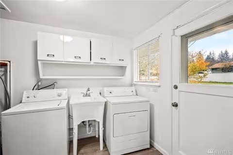 A bright laundry room with a white washing machine and dryer side by side, a white utility sink in between them, white cabinets mounted on the wall above, a window with blinds letting in natural light, and a door with a window showing an outdoor view.