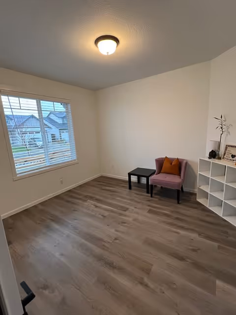 A small, minimally furnished room with light-colored walls and wood flooring. There is a window with white blinds on the left wall, a ceiling light fixture, a pink upholstered chair with an orange pillow, a small black side table, and a white cubical shelving unit with decorative items on the right side.