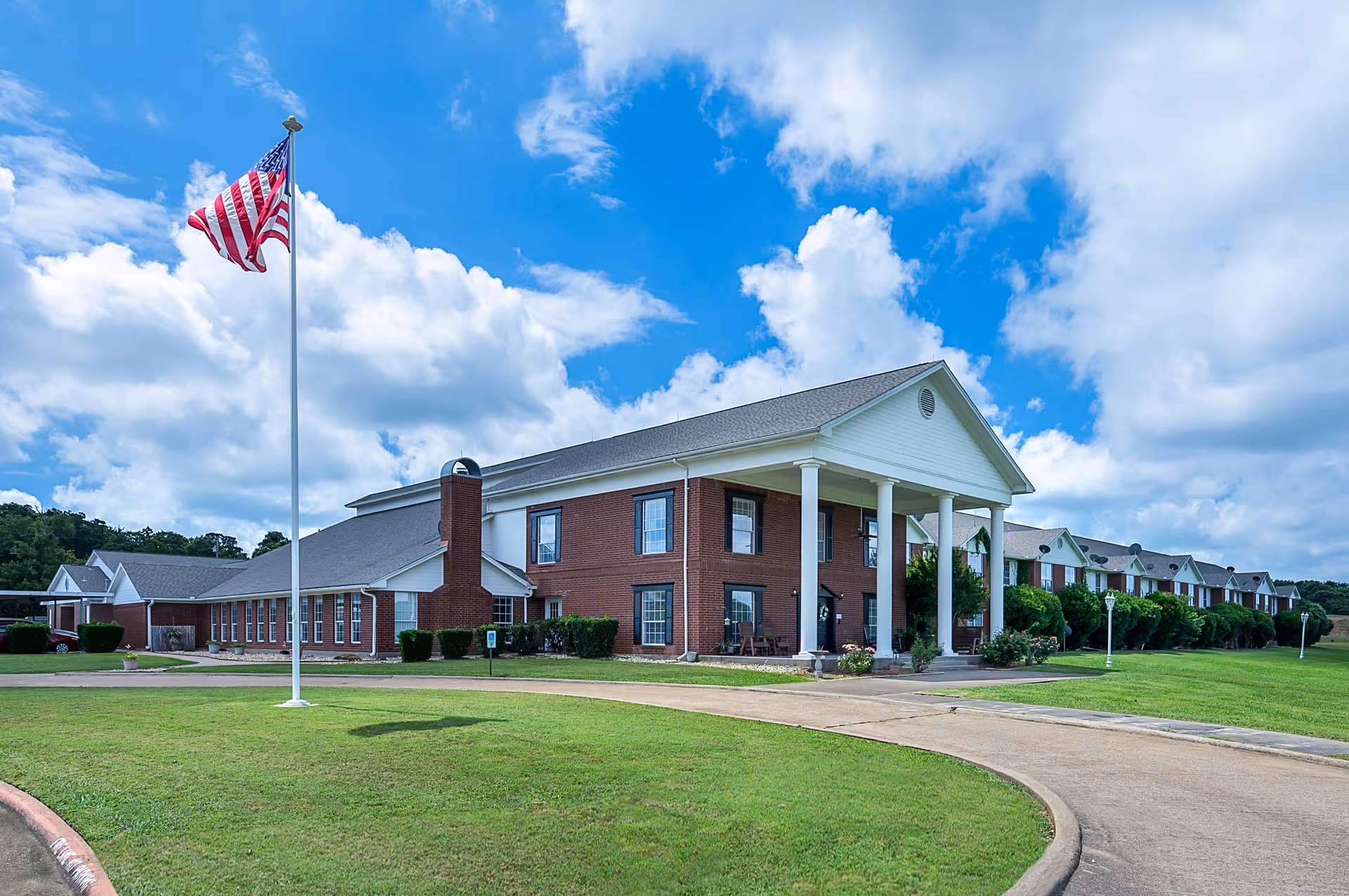 Exterior view of Pegasus Landing of Chisholm Trail, a large brick building with white columns at the entrance, surrounded by green grass and a curved driveway. An American flag on a tall flagpole is prominently displayed in the foreground under a partly cloudy blue sky.