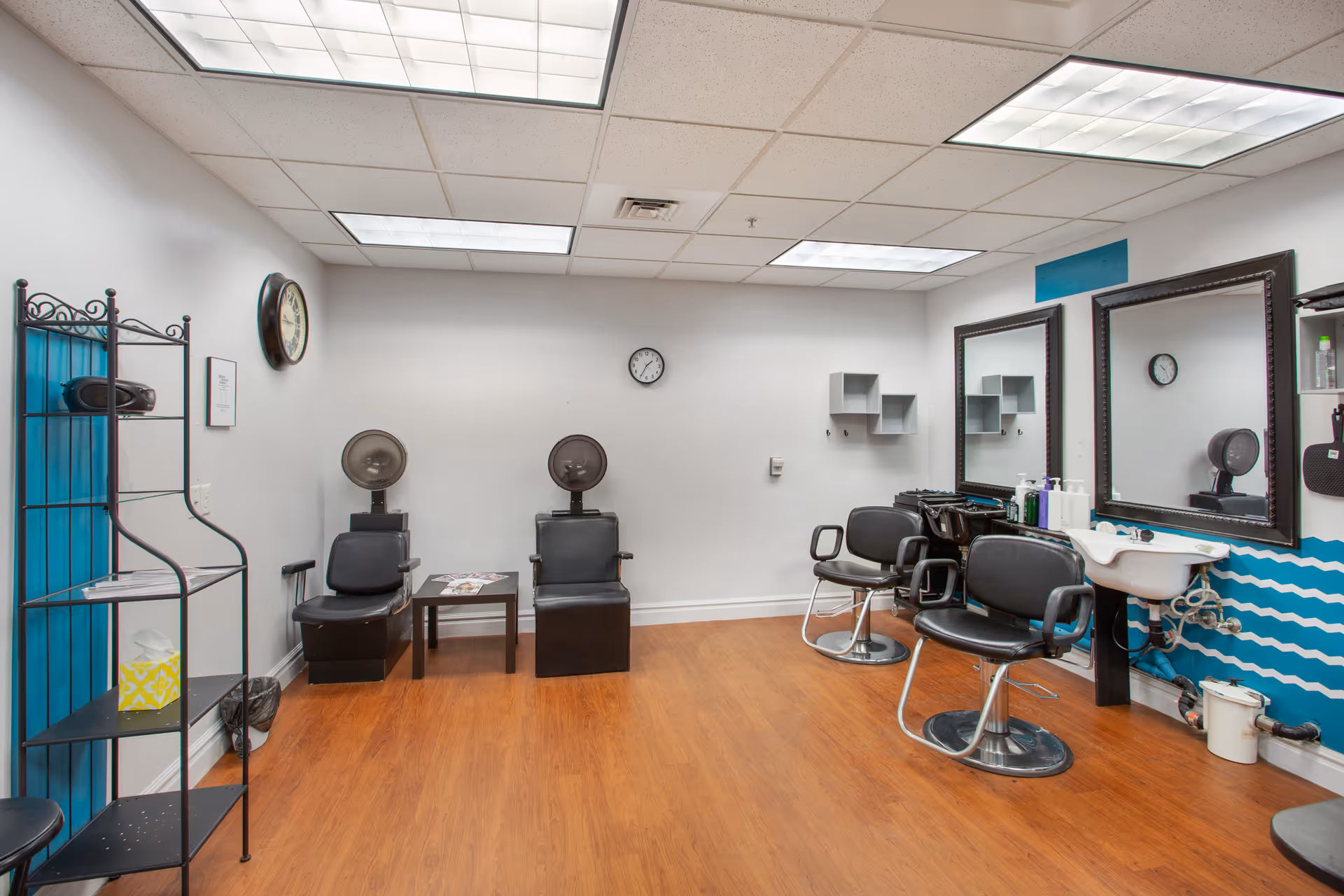 Interior of a hair salon with two black salon chairs in front of mirrors and a white sink. Two black hair dryer chairs are positioned against the back wall with a small table between them. The floor is wooden, and the walls are white with a blue accent featuring a wavy pattern. A black metal shelf stands on the left side of the room, and two clocks are mounted on the walls.