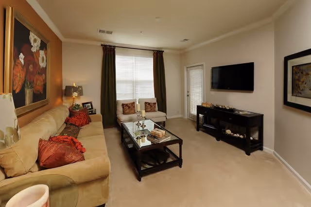 A cozy living room with a beige sofa adorned with red and brown pillows, a glass coffee table with books and candles, a small loveseat near a window with dark curtains, a flat-screen TV mounted on the wall above a black media console, and framed artwork on the walls.