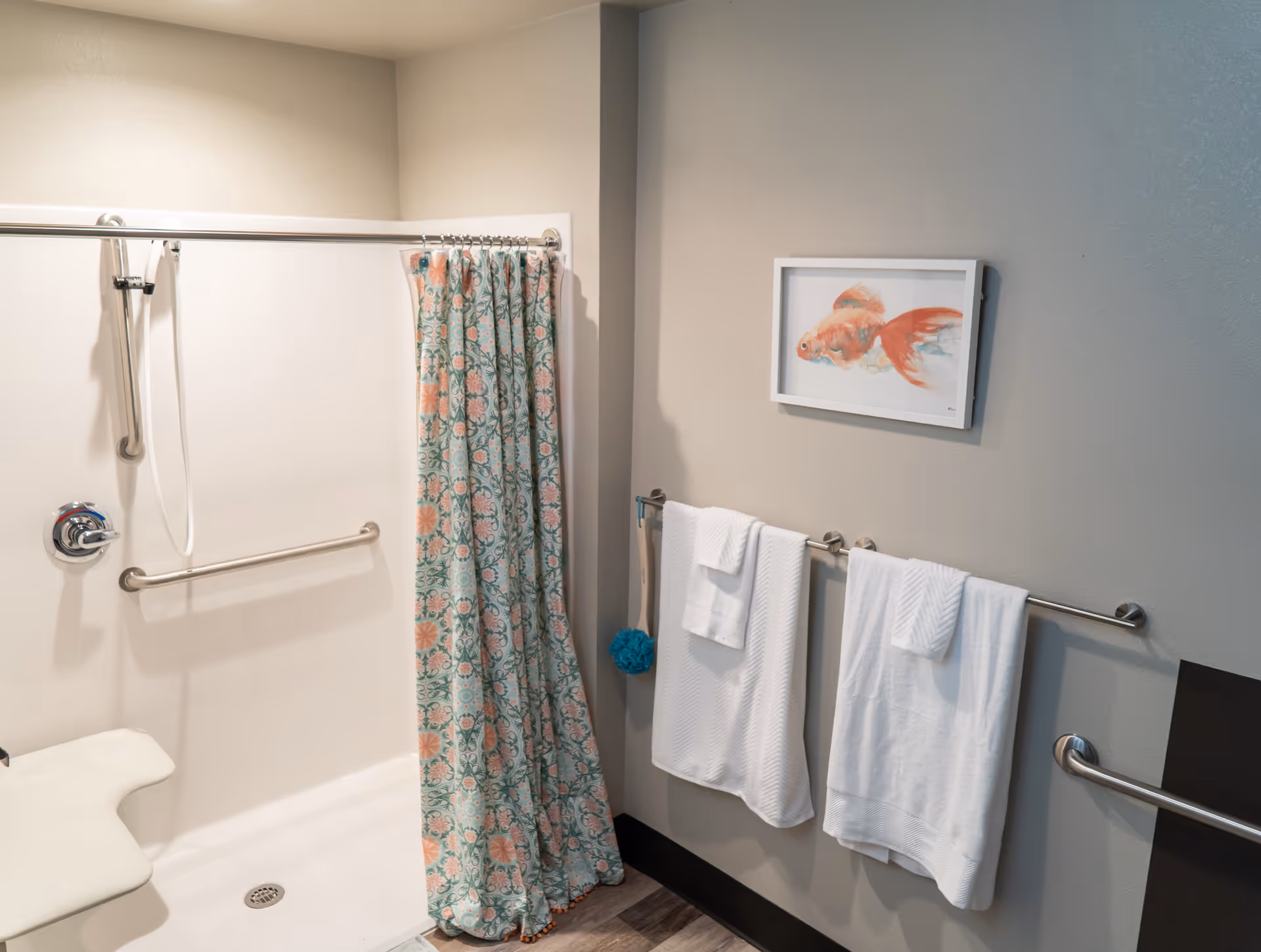A bathroom with a walk-in shower featuring a floral patterned shower curtain, a white shower seat, and grab bars. Two white towels hang on a towel rack on the wall, which also has a framed picture of a goldfish. The floor has wood-like tiles.
