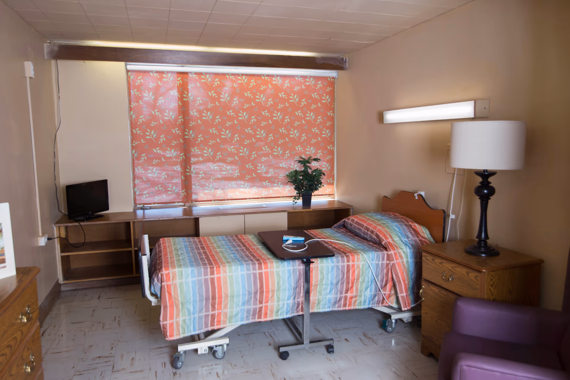 A small, simple bedroom in a rehabilitation center featuring a single hospital-style bed with a colorful striped bedspread. The room has a wooden nightstand with a lamp, a small TV on a low shelf, a purple armchair, and a window with floral-patterned blinds. There is also a small overbed table with a medical device on it.