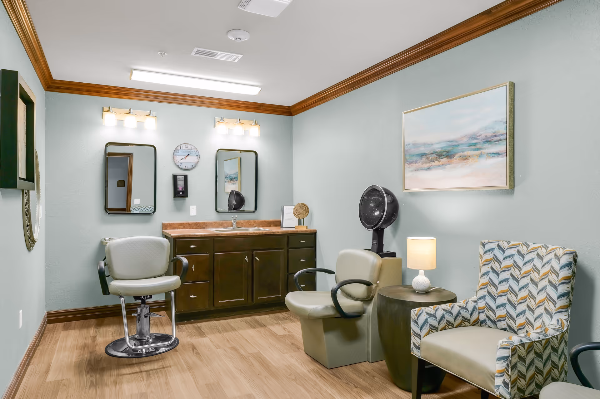 Interior of a salon room in a senior living facility featuring a styling chair, a sink with a countertop and cabinets, two mirrors with lights above them, a clock on the wall, a hair dryer chair, a side table with a lamp, and a patterned armchair. The walls are painted light green with wood trim and the floor is wood.