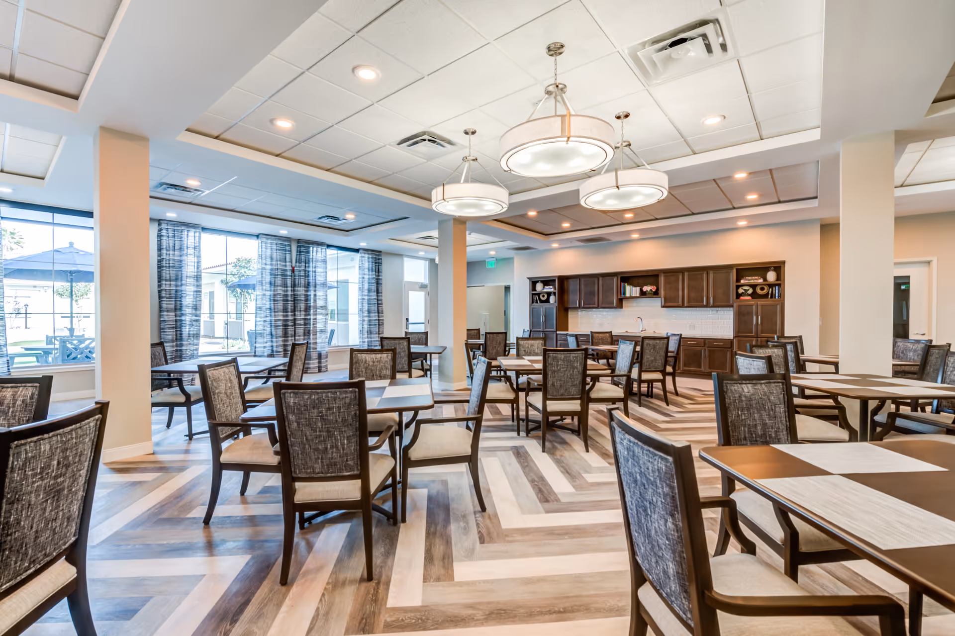 Bright communal dining room with multiple tables and chairs, large windows, and a kitchenette/serving area in the back.