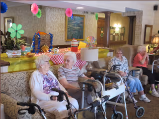 Four elderly women sitting in a common area with walkers in front of them. The room is decorated with colorful paper decorations and a table with party supplies and snacks in the background.