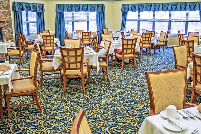 Dining room with multiple round tables set with white tablecloths and wooden chairs, patterned carpet, and large windows with blue valances.