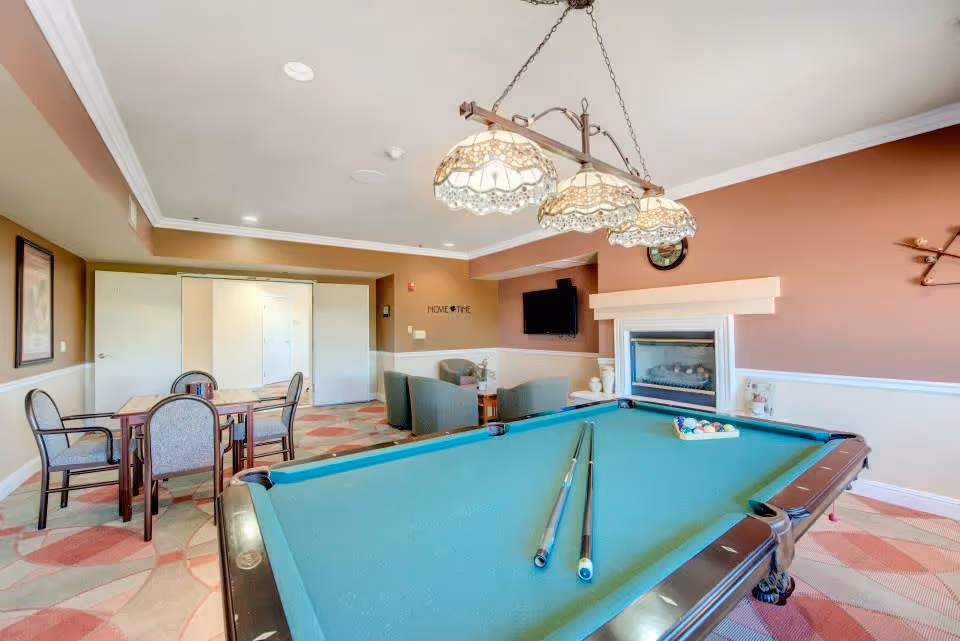 A recreational room with a pool table in the foreground, two pool cues resting on it, and a rack of billiard balls. In the background, there is a seating area with four chairs around a small table, a wall-mounted TV above a fireplace, and a dining table with four chairs. The room has beige and brown walls, a patterned carpet, and a hanging light fixture above the pool table.
