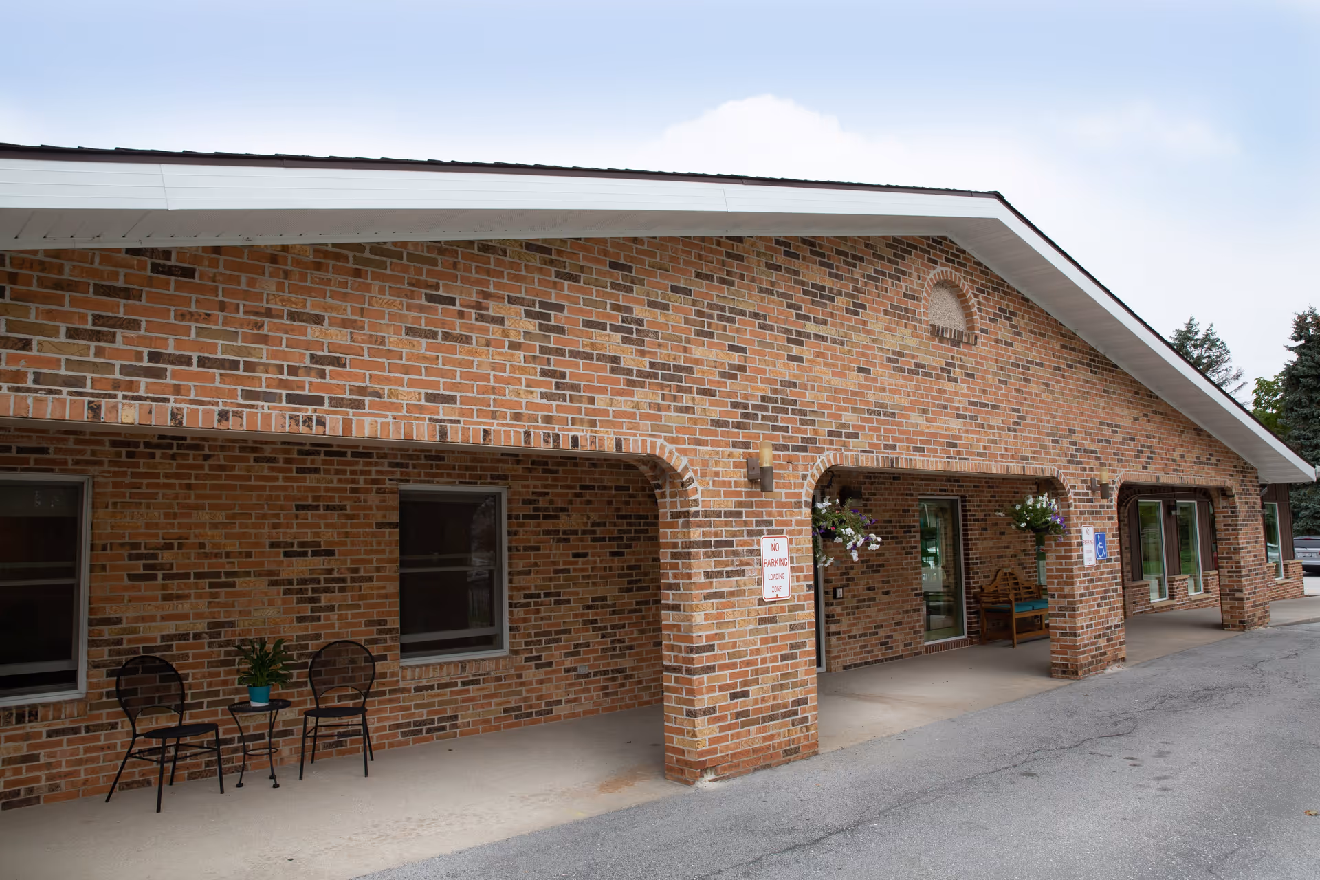 Brick building front with a covered walkway, chairs and hanging flower baskets by the entrance.