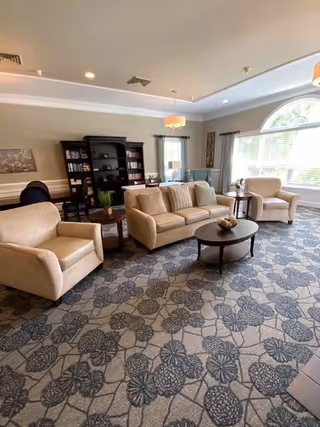A bright assisted-living common room with beige sofas and armchairs arranged around coffee tables, a bookshelf, and a large arched window.