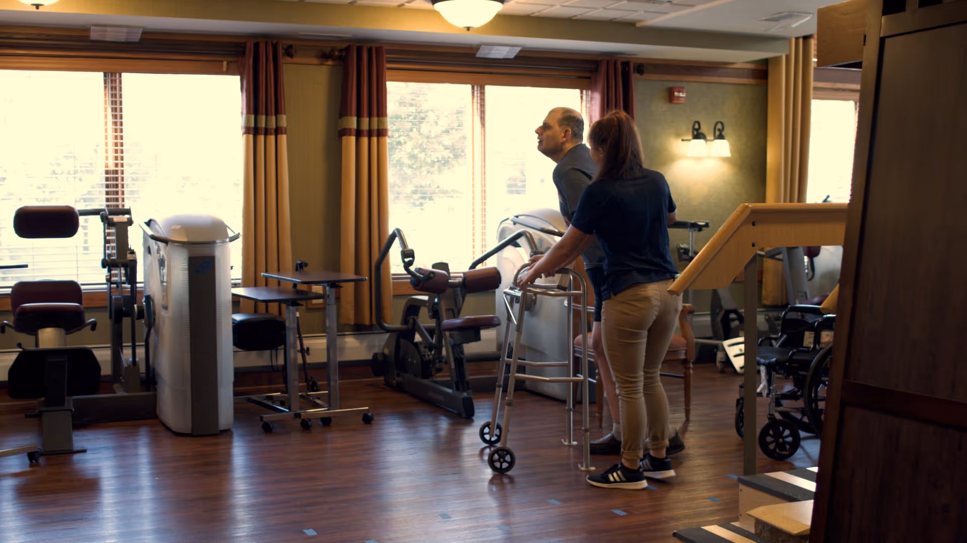 A rehabilitation room with exercise equipment and large windows with curtains. A man using a walker is assisted by a woman standing beside him. The room has wooden flooring and warm lighting.