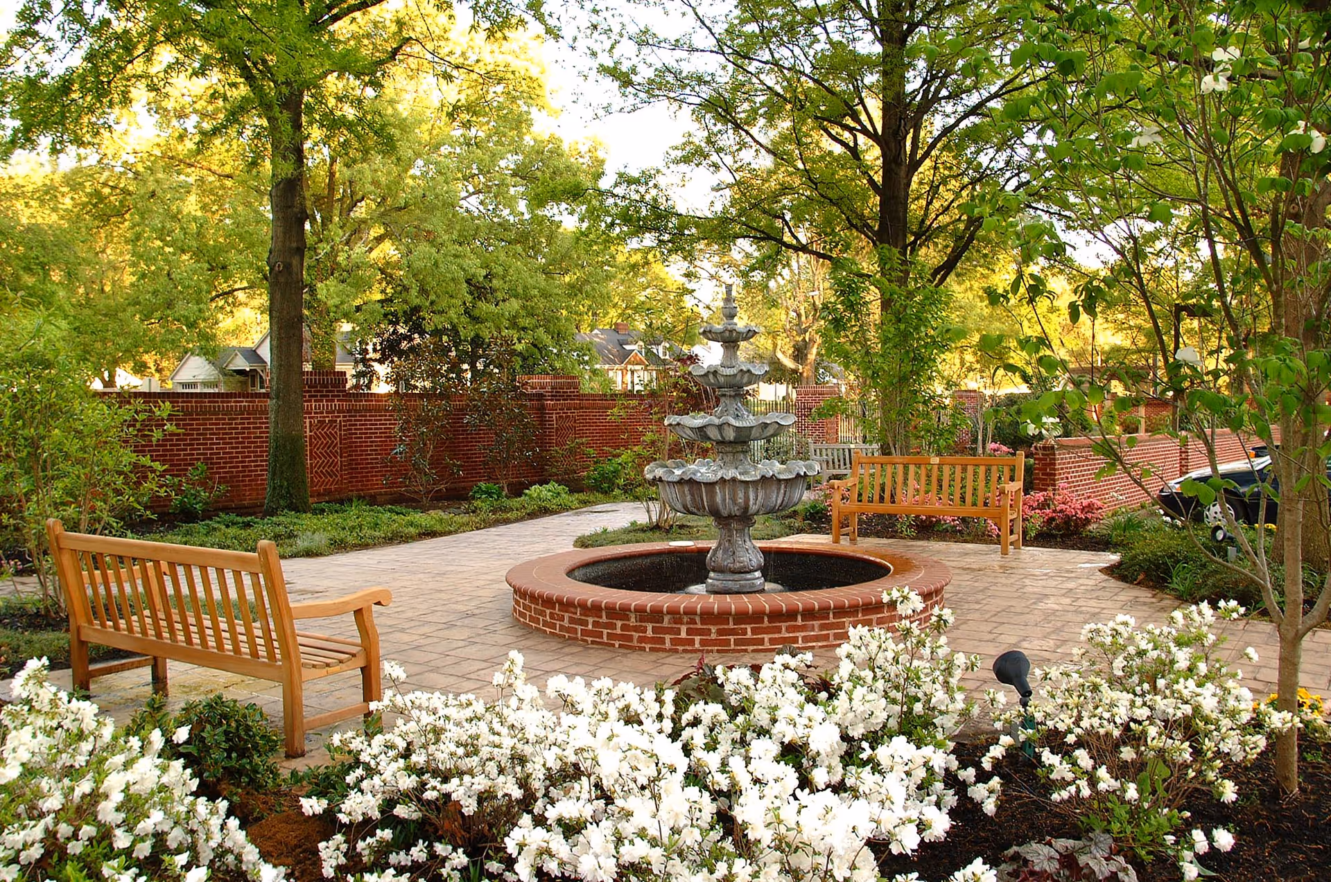 A peaceful outdoor garden area with a multi-tiered stone fountain in the center, surrounded by a circular brick border. There are wooden benches placed around the fountain on a paved patio. The garden is lush with green trees, bushes, and white flowering plants, enclosed by a red brick wall.