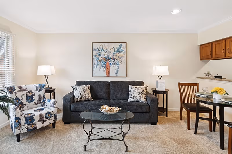 A cozy living room with a dark gray sofa adorned with two patterned pillows, a glass coffee table with decorative items, a floral-patterned armchair, two side tables each holding a lamp, a framed floral painting on the wall, and a small dining area with a round glass table and two wooden chairs. The room has beige carpet and light-colored walls.