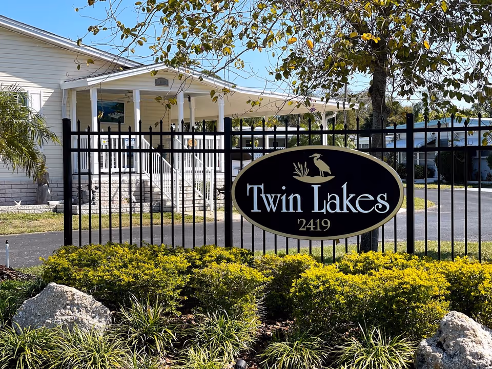 Black metal fence with an oval sign reading 'Twin Lakes 2419' in front of a white building with a porch and stairs, surrounded by green bushes and trees under a clear blue sky.