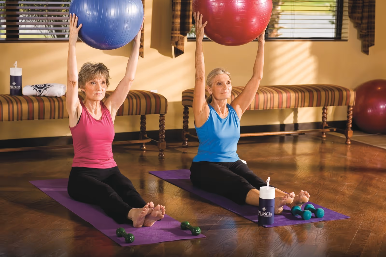 Two older women sitting on purple exercise mats in a room with wooden floors, holding large exercise balls above their heads. Each woman has a pair of small green dumbbells and a water bottle next to her. The room has striped benches and windows with blinds.