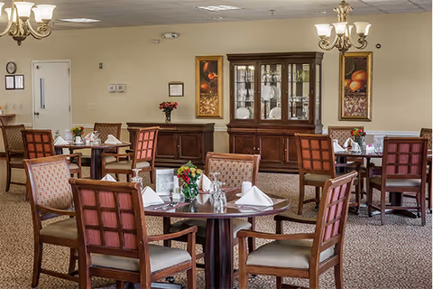 Dining room with round tables set for meals, wooden chairs, chandeliers, floral centerpieces, and a china cabinet against the wall.