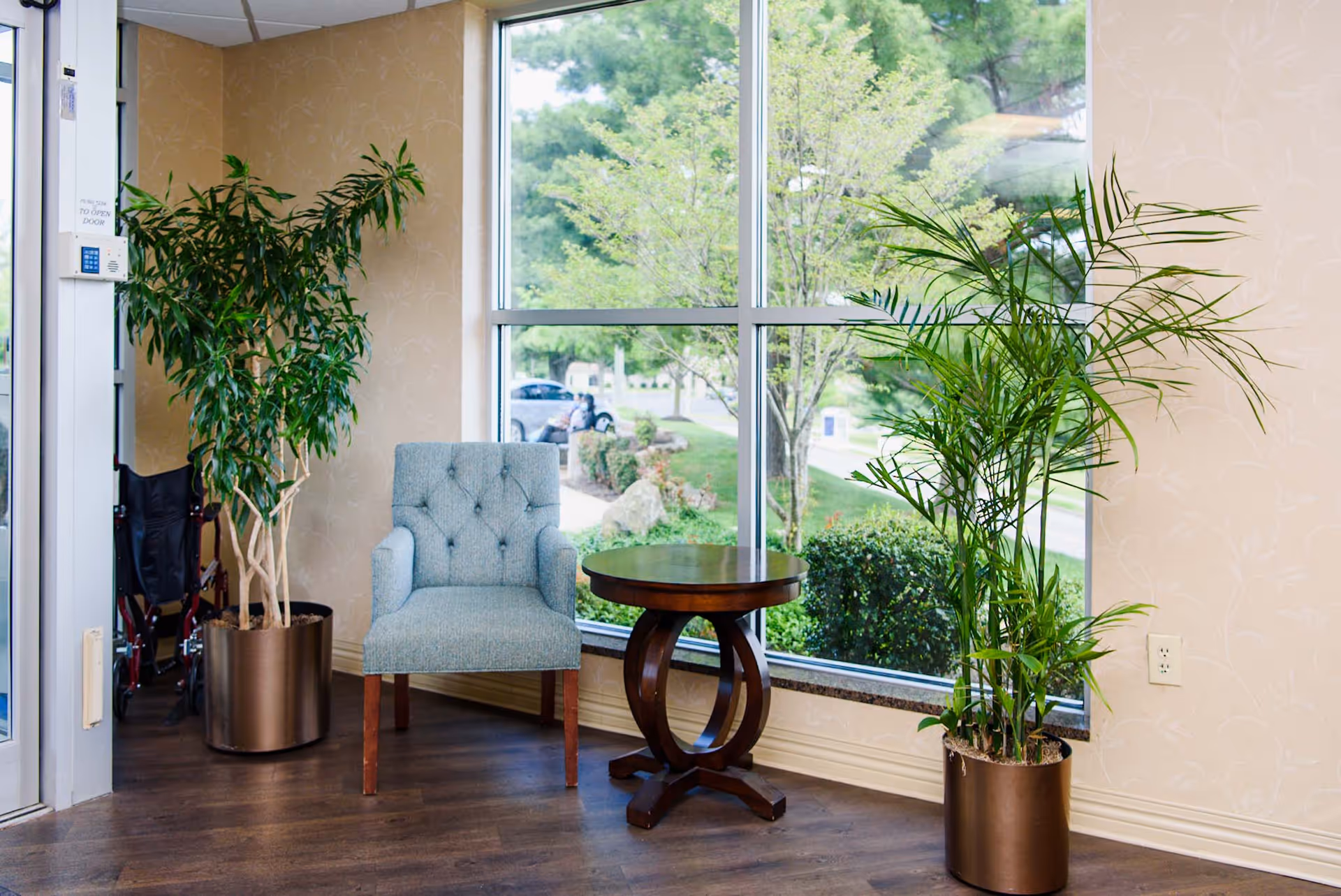 A cozy corner inside a facility with a light blue upholstered armchair and a round wooden side table placed near a large window. Two tall potted plants flank the chair, and outside the window, greenery and a parked car are visible. The floor is wooden, and the walls have a light beige wallpaper with a subtle pattern.