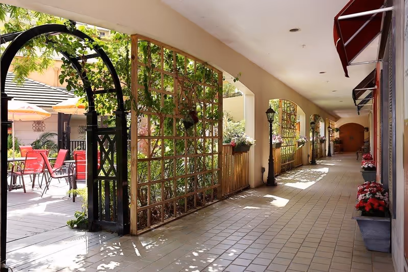 A bright, tiled corridor with arched openings on one side featuring wooden trellises with green plants and flowers. On the left, there is an outdoor patio area with red chairs and tables under umbrellas. The corridor is decorated with flower pots and vintage-style lamp posts.