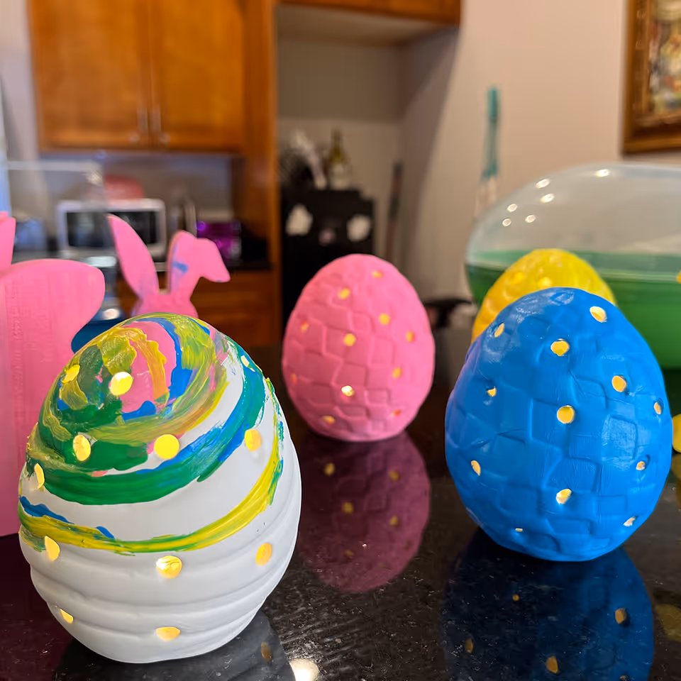 Close-up of colorful decorative Easter eggs with small holes, placed on a shiny black countertop in a kitchen setting. In the background, wooden cabinets, a microwave, and other kitchen items are visible.