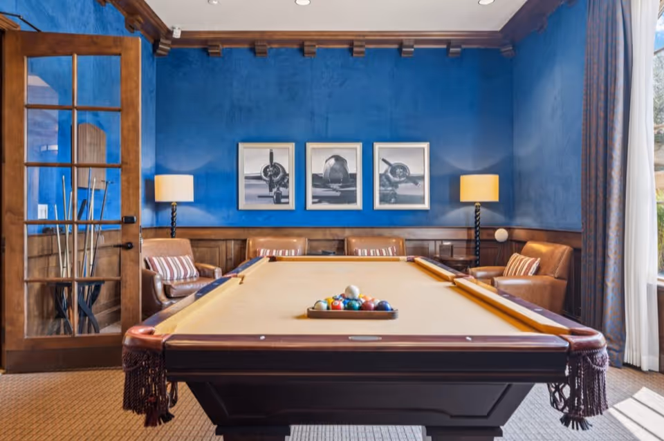 A cozy game room with a pool table in the center, featuring a beige felt surface and a rack of billiard balls. The room has blue walls with three black and white airplane pictures hanging above a wooden wainscoting. There are two leather armchairs with striped cushions on either side of the pool table, each accompanied by a floor lamp. A glass-paneled wooden door is on the left, and a window with curtains is on the right, allowing natural light to enter.