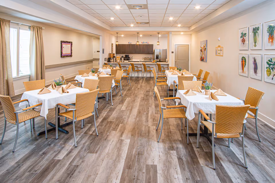 Dining room with several tables covered in white tablecloths, wicker chairs, and a serving counter/kitchen area at the back.