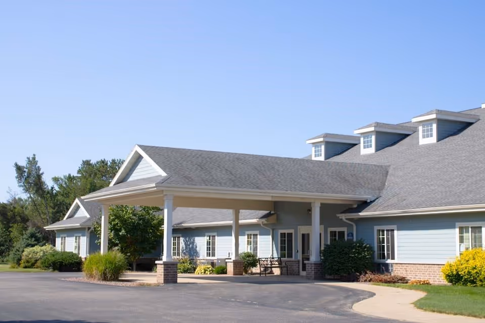 Exterior view of a single-story senior living facility building with a covered entrance supported by white columns. The building has light blue siding, multiple windows, and a gray shingled roof with dormer windows. There are bushes and greenery around the building and a clear blue sky above.