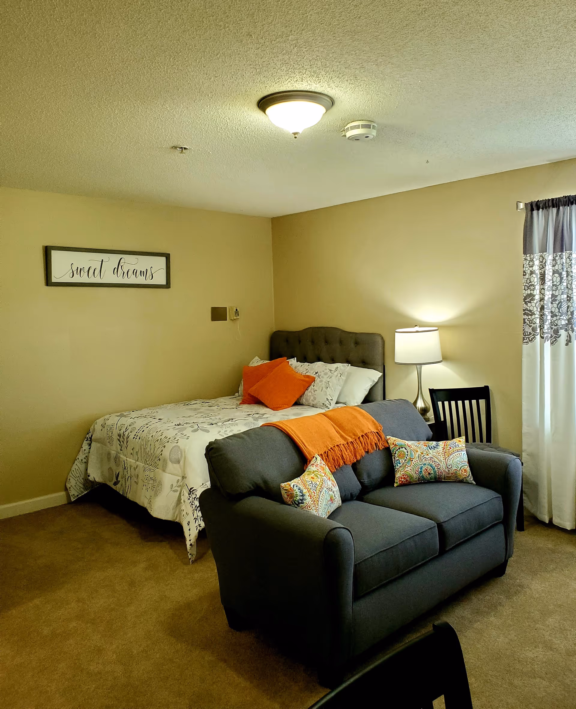 A cozy bedroom with a bed against the wall, decorated with white and gray bedding and orange pillows. In front of the bed is a dark gray loveseat with colorful patterned pillows and an orange throw blanket. A lamp on a small table and a chair are next to the bed. A window with white and gray patterned curtains is on the right side. A framed sign on the wall above the bed reads 'sweet dreams.'