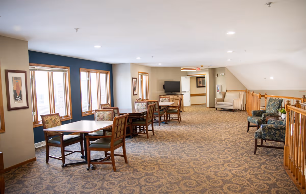 A spacious common area in a senior living facility with several tables and chairs arranged near windows on the left side. The room features patterned carpet, beige walls with a dark blue accent wall, framed artwork, and a TV mounted on a cabinet in the background. There are additional seating options including armchairs along the right side near a wooden railing.