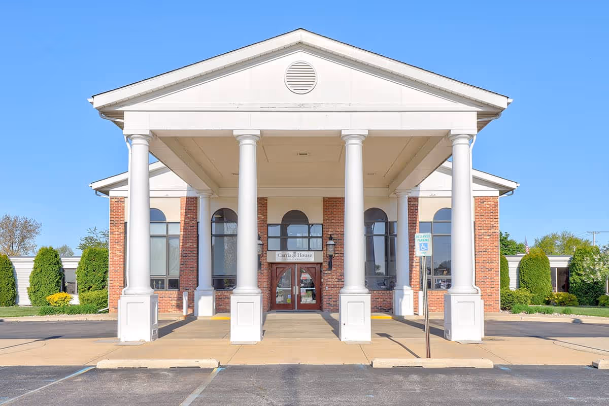 Front exterior view of Carriage House Nursing and Rehab building with large white columns supporting a covered entrance, brick walls, large windows, and a clear blue sky.