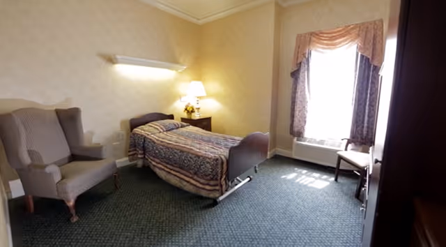 A small bedroom with a single bed covered in a patterned bedspread, a wooden nightstand with a lamp and a small flower arrangement, a cushioned armchair, a window with draped curtains, and a chair near the window. The room has carpeted flooring and light-colored walls.