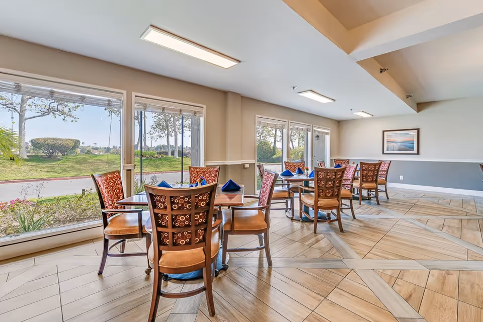 A bright dining room with several wooden tables and chairs arranged neatly. Each table has blue folded napkins. Large windows provide a view of a green outdoor area with trees and bushes. The room has light-colored walls, a framed picture on one wall, and a patterned tile floor.