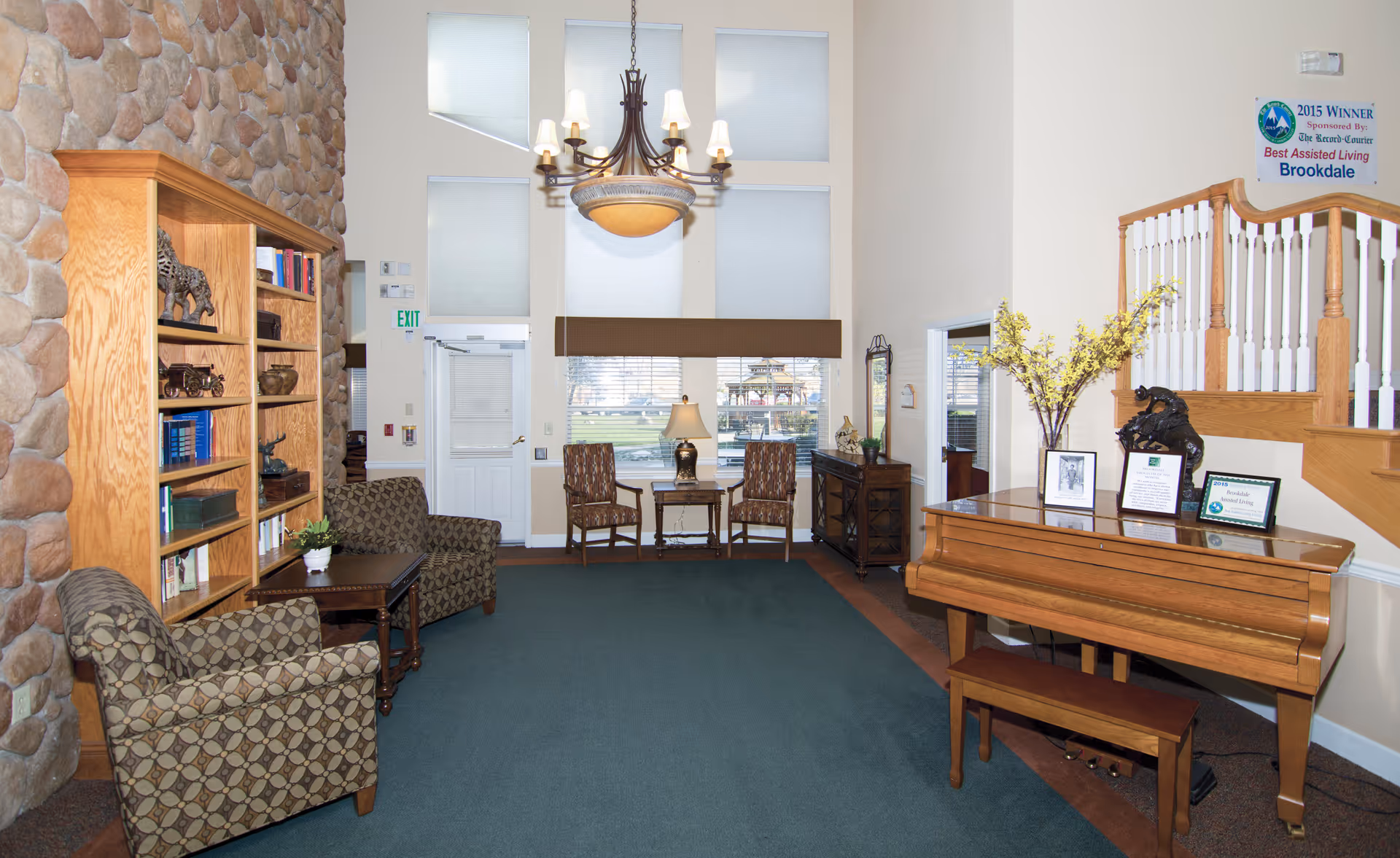 A cozy living room area with a stone wall on the left, wooden bookshelf filled with books and decorative items, two patterned armchairs with a small wooden table between them, a large window with blinds and a brown valance, two wooden chairs with a table and lamp between them, a wooden piano with a bench on the right, and a staircase with white spindles and wooden railing. There are framed awards and a vase with yellow flowers on the piano. A chandelier with multiple lights hangs from the ceiling.