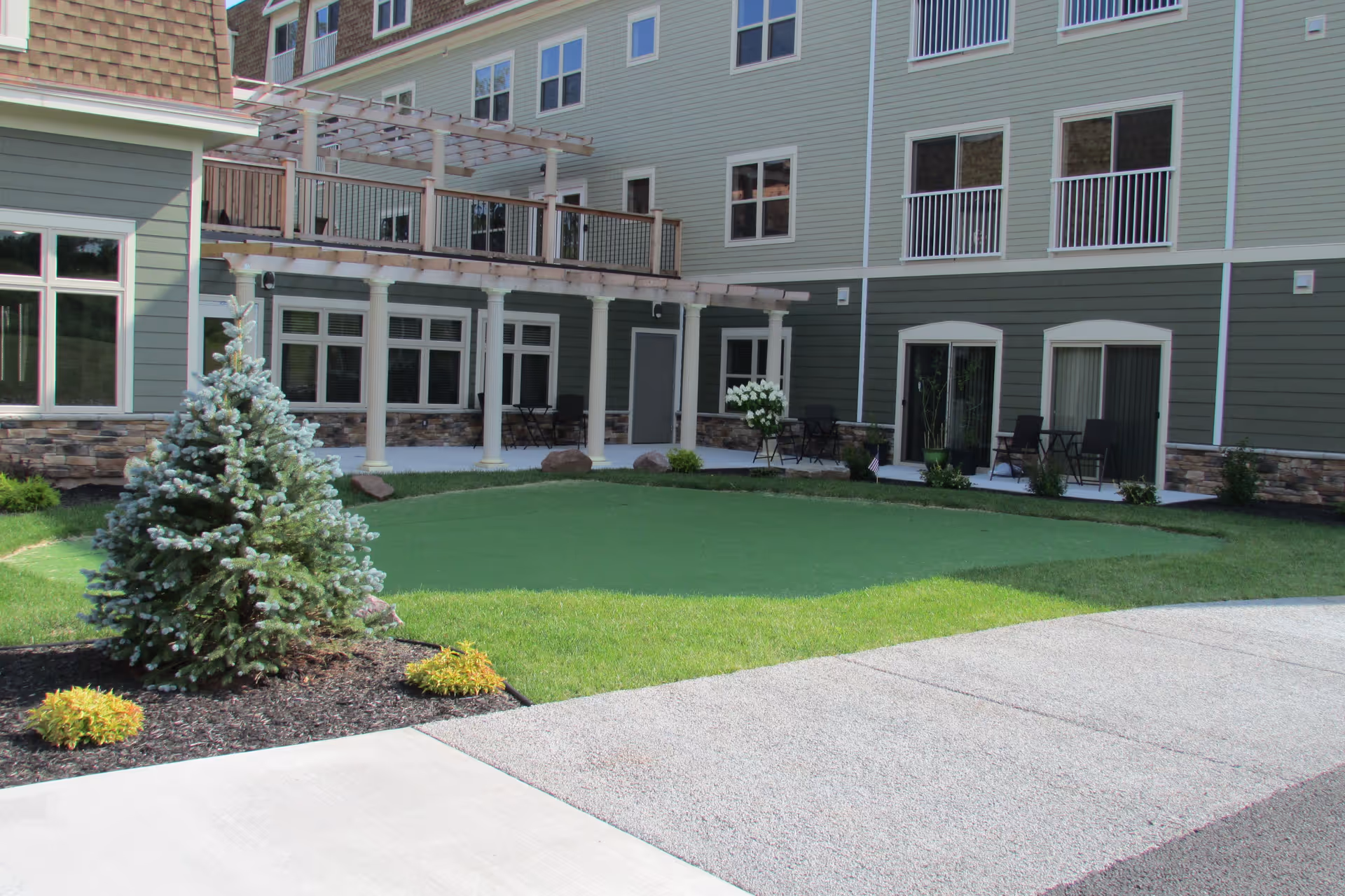 Outdoor courtyard area of a senior living facility with a small artificial putting green, a young pine tree, and several small plants. The building has multiple windows and balconies, with a covered patio area supported by white columns and furnished with chairs and tables.