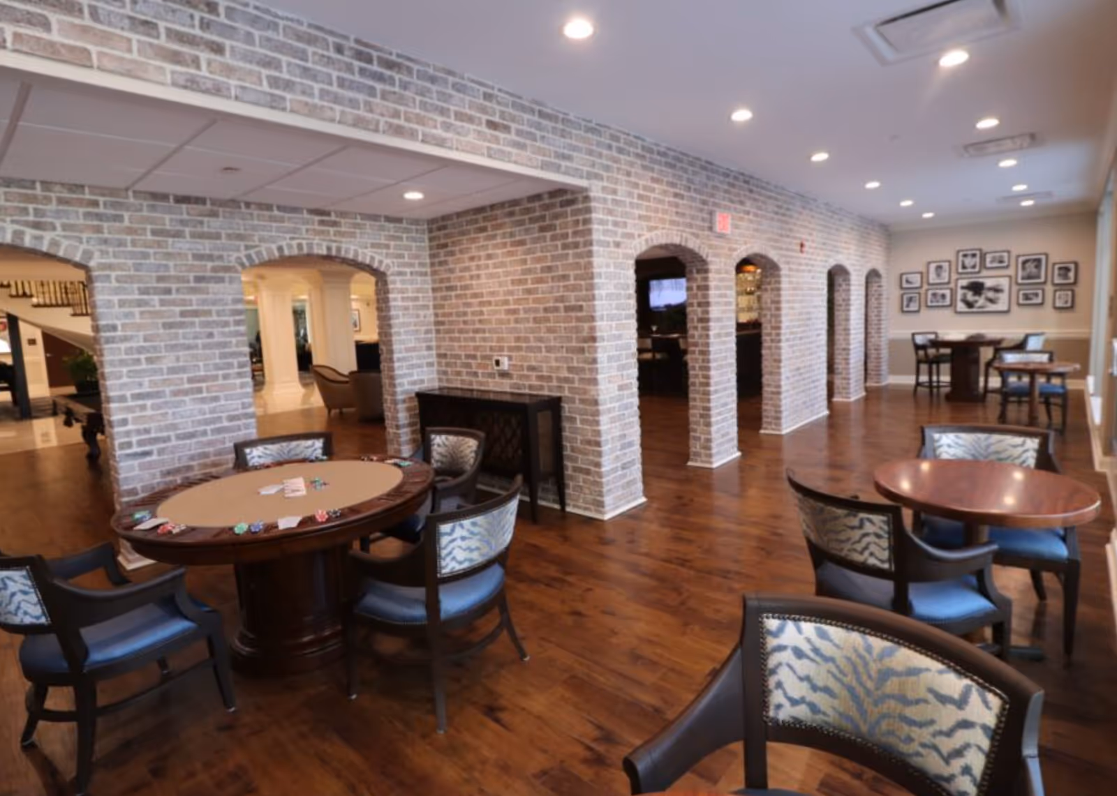 A spacious senior living common room with round card and dining tables, chairs, and exposed brick archways.