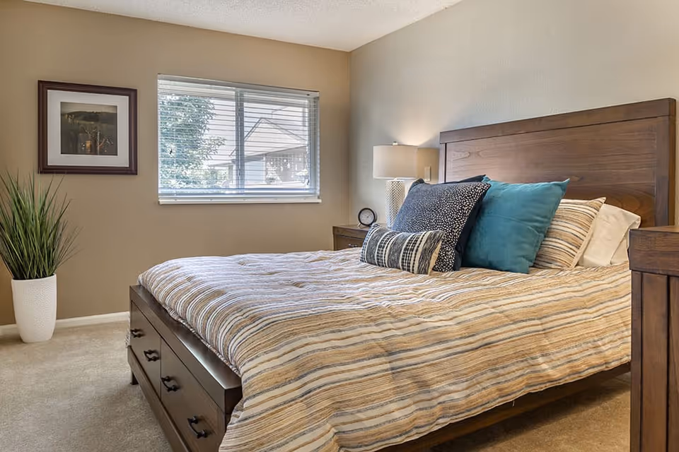 Well-lit bedroom with a wooden bed dressed in striped bedding and decorative pillows, a nightstand with a lamp, a window, framed art, and a potted plant.