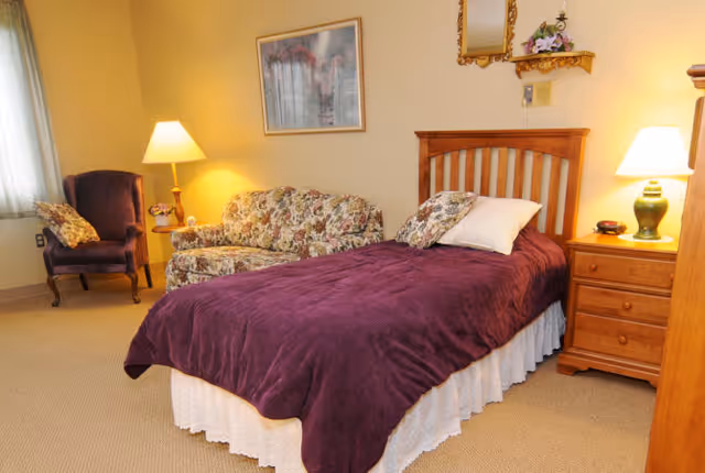 A cozy bedroom with a single bed covered in a purple blanket and white bed skirt. Next to the bed is a wooden nightstand with a green lamp and a clock. There is a floral patterned loveseat and a purple armchair near a window with sheer curtains. The walls are painted light yellow, decorated with a framed picture and a small shelf with a mirror and flowers. The room is warmly lit by two lamps.
