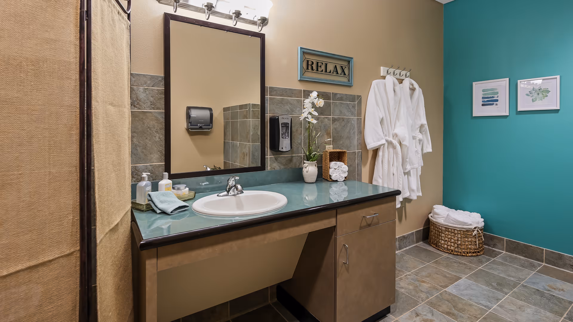 A clean and well-lit bathroom with a green countertop sink, a large mirror, and a wall-mounted soap dispenser. Two white bathrobes hang on hooks on a beige wall next to a basket filled with towels. The wall on the right is painted teal and decorated with two framed pictures. A sign above the sink reads 'RELAX'.