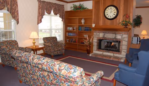 A cozy living room with floral patterned sofas and two blue armchairs arranged around a stone fireplace with a wooden mantel. Above the fireplace is a large clock. There are built-in wooden bookshelves with books and decorative items, two windows with floral valances, and two table lamps providing warm lighting.