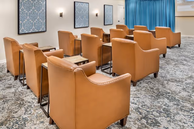 A small theater or presentation room with two rows of tan leather armchairs arranged facing a screen. Each chair has a small wooden side table next to it. The walls are decorated with three framed blue patterned artworks and two wall sconces. Blue curtains cover the front wall near the screen.