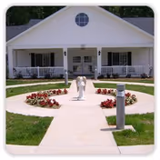 Front entrance of a single-story white building with a circular walkway, central statue, and flower beds leading to a covered porch.