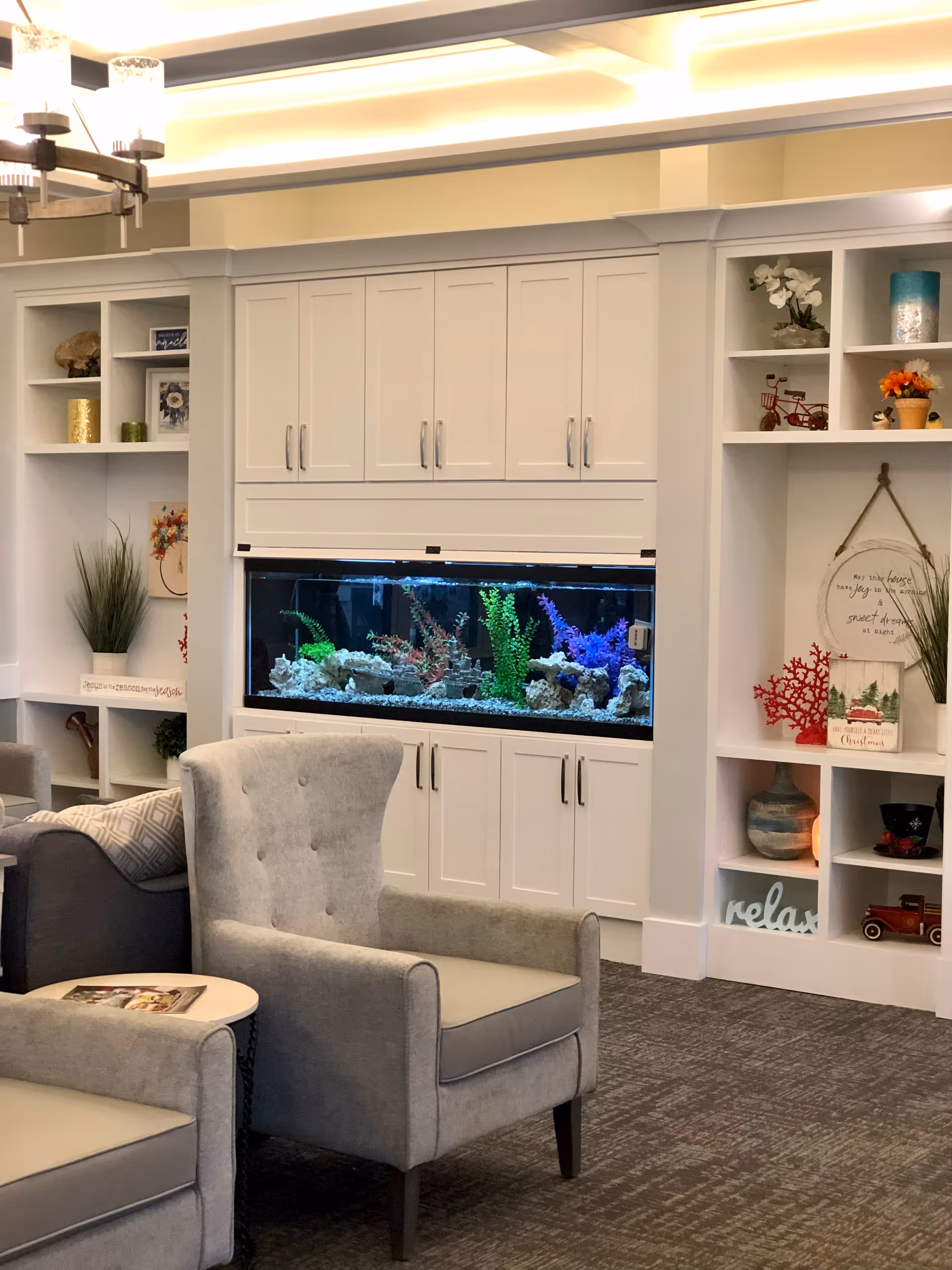 A cozy living room area with two gray upholstered armchairs and a small round table. Behind the chairs is a built-in white cabinet with an aquarium filled with colorful aquatic plants. The surrounding shelves are decorated with various ornaments, plants, and framed pictures. The room has soft lighting and a carpeted floor.