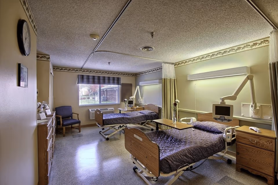A two-bed patient room in a senior healthcare facility with hospital beds, over-bed tables, medical monitors, and a window.