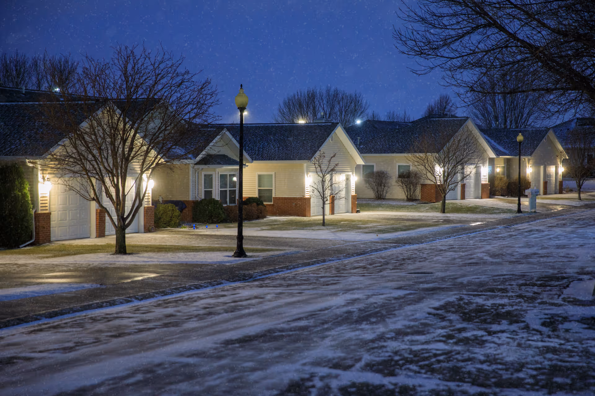 Row of single-story residential units with lit porch lights and street lamps on a snowy evening.