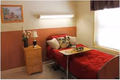 Small private bedroom with a single bed dressed in red bedding, an overbed tray, a bedside cabinet with a lamp and flowers, and a window.