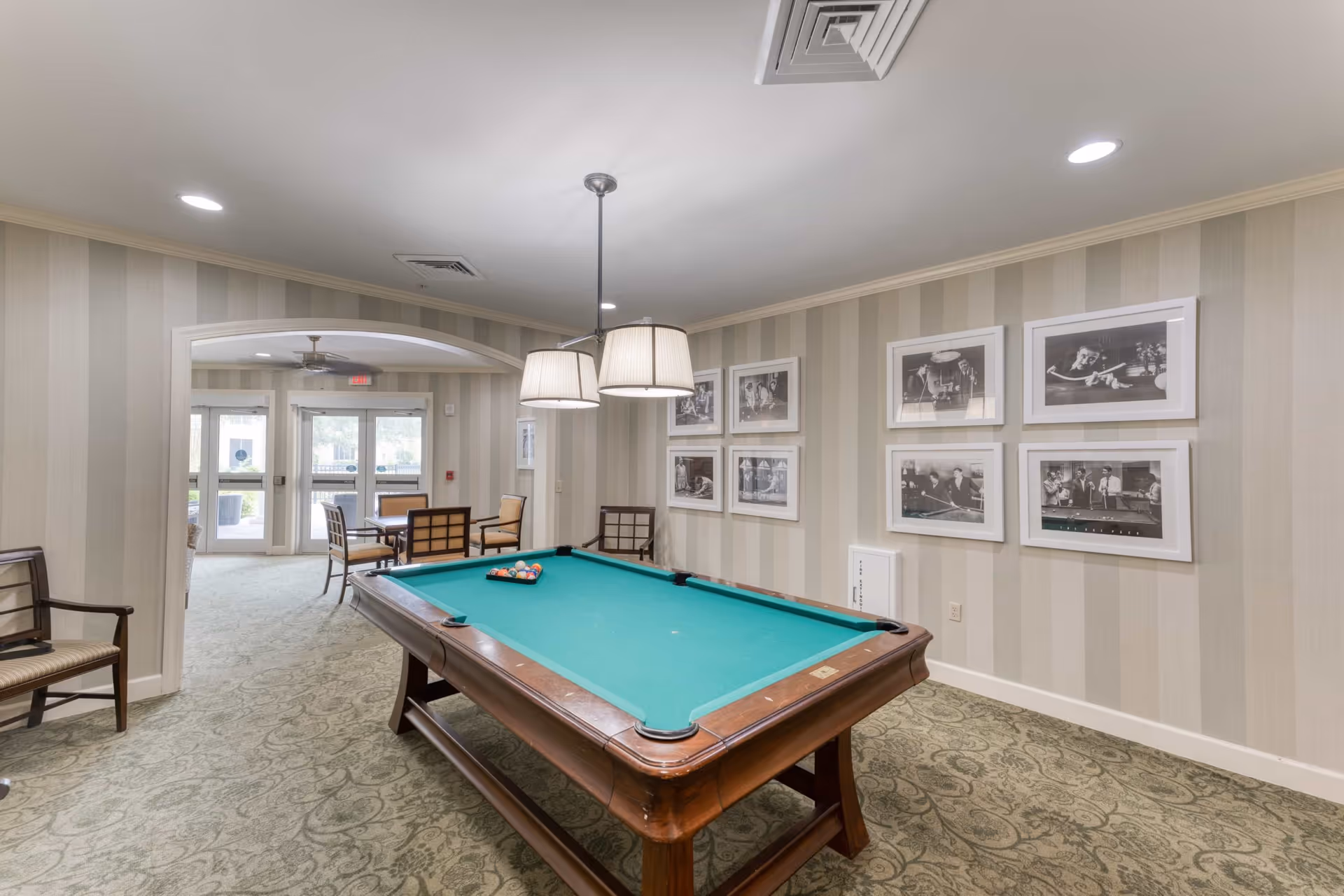 A recreational room with a pool table in the center, surrounded by chairs. The walls have striped wallpaper and are decorated with framed black and white photos. There is a hanging light fixture above the pool table and an adjacent seating area with a table and chairs near glass doors leading outside.
