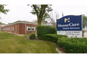 Exterior view of a single-story brick building with windows, surrounded by grass and bushes. A large blue and white sign in front reads 'ManorCare Health Services Nursing & Rehab MedBridge Unit' with an emblem above the text.