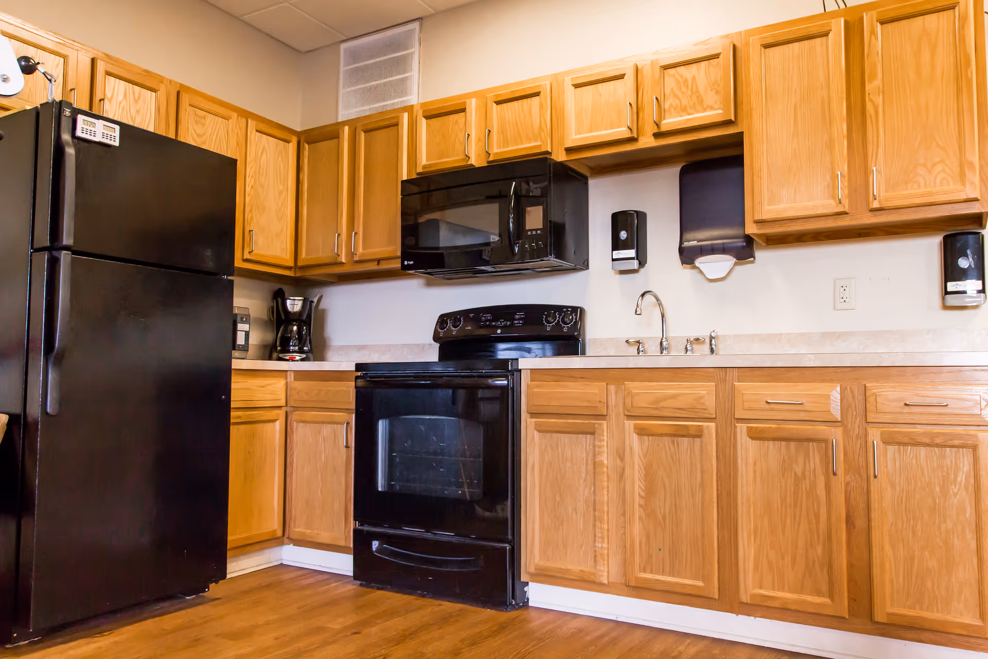 A kitchen area with wooden cabinets, a black refrigerator, black microwave, black stove, a coffee maker, and a sink with a faucet. The countertop is light-colored, and there are wall-mounted soap and paper towel dispensers above the sink. The floor is wooden.