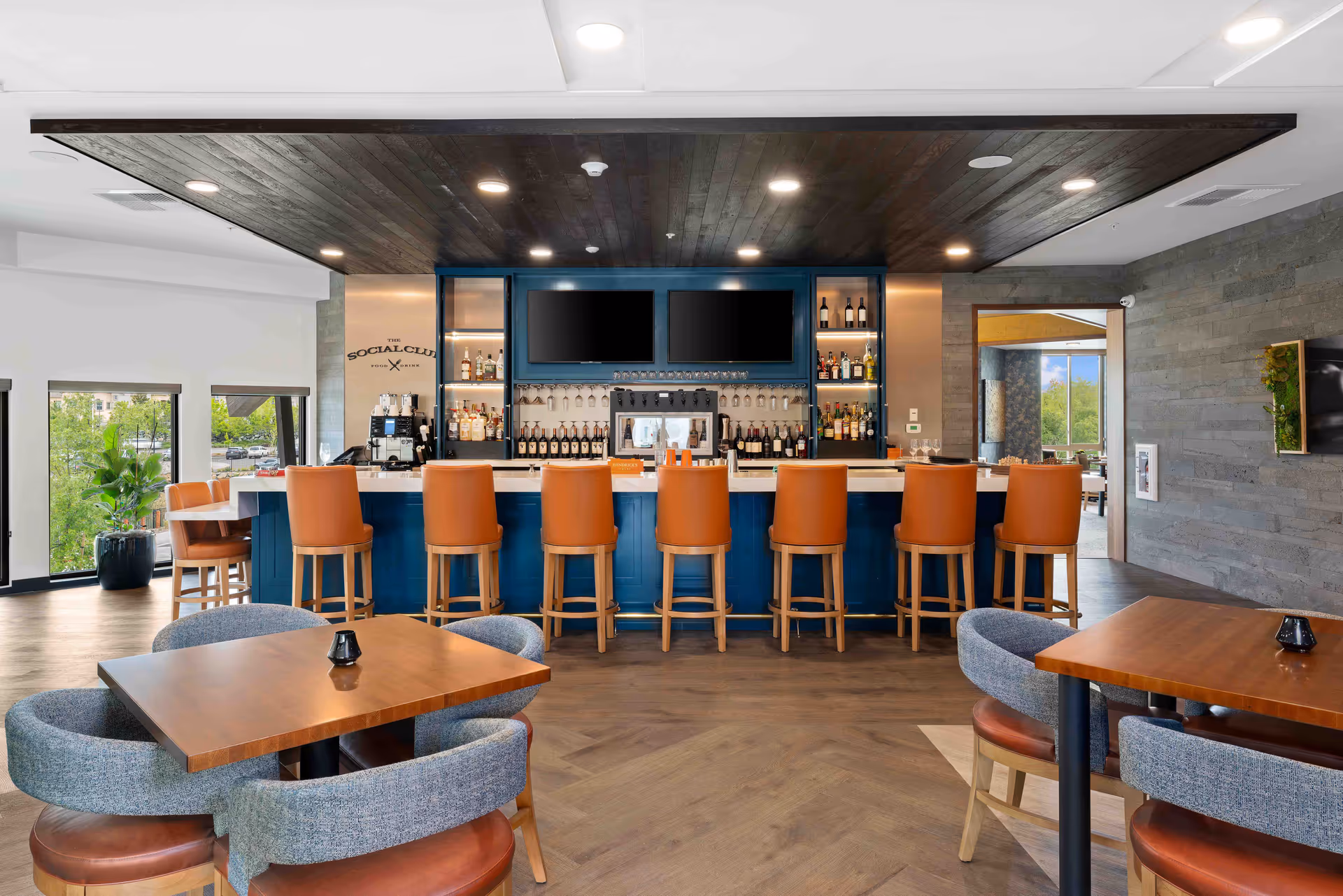 Interior view of a modern bar area with a blue bar counter and eight brown leather bar stools. Behind the bar are shelves stocked with bottles of liquor and two large flat-screen TVs mounted on the wall. In the foreground, there are wooden tables with cushioned chairs. Large windows on the left side provide natural light and a view of greenery outside.